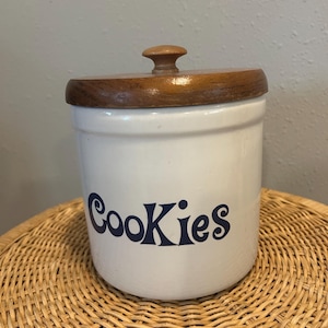 May include: A cylindrical white ceramic cookie jar featuring the word Cookies printed in dark blue script lettering on the front. The jar is topped with a circular brown wooden lid that includes a small, rounded wooden knob handle at the center.