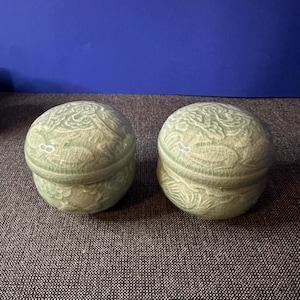 May include: Two celadon green ceramic lidded jars with a crackle glaze. The jars have a rounded shape and are decorated with a raised floral pattern. The jars are sitting on a gray textured surface.