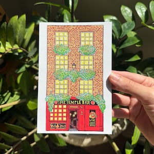 May include: A watercolor illustration of The Temple Bar in Dublin, Ireland. The building features a red facade with the bar's name and a sign that says "Live Music Daily." The artwork includes brickwork, windows, and green foliage.