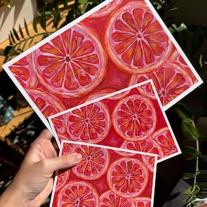 May include: Three art prints featuring a vibrant watercolor illustration of blood orange slices. The prints vary in size, showcasing the fruit's red and orange hues with white borders. The artwork is displayed against a backdrop of green foliage.