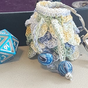 May include: A crocheted dice bag with a blue, green, and yellow color scheme. The bag has a silver metal clasp and two blue and white swirl beads. A blue and silver metal dice is in the background.
