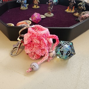 May include: A pink crocheted dice bag with a silver keychain and a blue iridescent d20 dice.