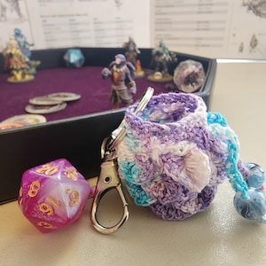 May include: A purple, white, and blue crocheted dice bag with a silver keychain and a pink and white iridescent 20-sided die.