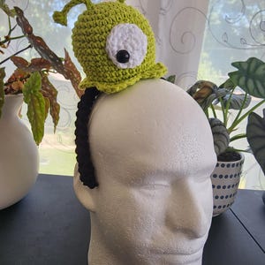 May include: A lime green crocheted alien hat with a large white eye and black pupil, attached to a black headband. The hat features two antennae and a ruffled brim. The hat is displayed on a white foam head, with plants in the background.