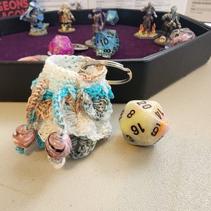 May include: A crocheted dice bag in shades of blue, white, and brown, with two decorative beads. A large, twenty-sided die sits next to the bag. The background includes miniature figurines and a black tray, suggesting a tabletop gaming theme.