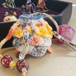 May include: A crocheted dice bag with a colorful pattern of blue, white, yellow, orange, and pink. The bag is attached to a silver keychain with a purple and gold dice charm.