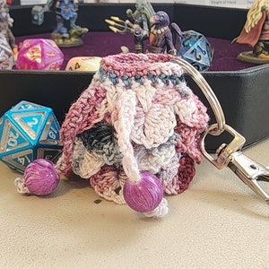 May include: A crocheted dice bag with a pink, white, and blue color scheme. The bag has a silver metal clasp and two purple beads. The bag is sitting on a white surface.