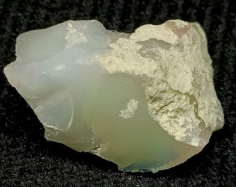 May include: A raw, unpolished opal gemstone with a translucent, milky white body and subtle iridescent flashes of color. The stone features a rough, textured, beige-colored matrix crust on one side, contrasting with the smooth, glassy surface.
