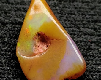 May include: A polished, teardrop-shaped opal gemstone. The stone features a mix of colors, including orange, yellow, and purple, with a small hole near the center. The opal is set against a dark, textured background.