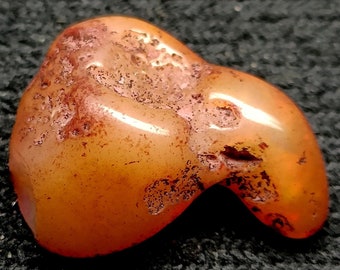 May include: A polished, orange-brown gemstone with a smooth, rounded shape and a textured, reddish-brown surface. The stone has a glossy finish and appears to be a natural, organic material. The gemstone is likely used for jewelry or decorative purposes.