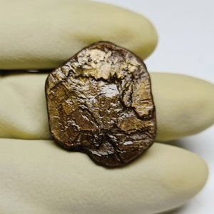 May include: A worn, copper-coloured coin with an irregular shape and a raised design. The coin has a dark patina and appears to be very old.