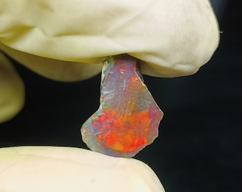 May include: A vibrant, translucent opal gemstone fragment. The stone displays a fiery mix of red, orange, and yellow hues, with hints of blue and purple. The opal is held by a gloved hand against a dark background.