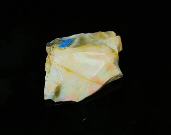 May include: A rough-cut, natural gemstone with a mix of colors. The stone features a creamy white base with streaks of yellow, pink, and green. A small patch of bright blue is visible on one edge. The stone is set against a black background.