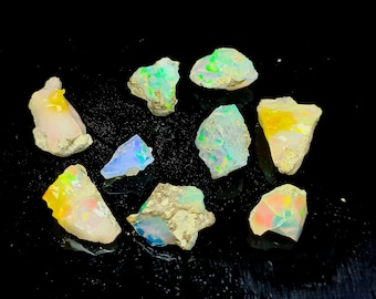 Puede incluir: Una colección de diez piedras preciosas de ópalo en bruto en varios tonos de blanco, azul, verde y amarillo. Los ópalos están dispuestos sobre una superficie negra.
