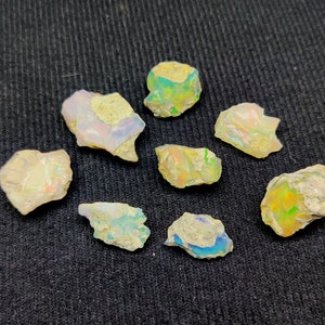 Puede incluir: Una colección de ocho pequeñas piedras de ópalo ásperas e iridiscentes en varios tonos de blanco, verde y amarillo. Las piedras están dispuestas sobre una superficie texturizada gris oscuro.