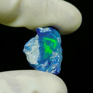 Puede incluir: Una piedra preciosa de ópalo azul y verde con una superficie rugosa y natural. El ópalo se sostiene en una mano enguantada.