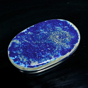 May include: A smooth, oval-shaped lapis lazuli cabochon with a deep blue color and white and gold flecks. The stone is polished and has a glossy finish.