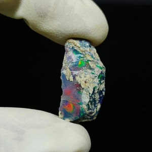 Puede incluir: Una piedra preciosa de ópalo en bruto con colores vibrantes. La piedra muestra una mezcla de azules, verdes y rojos, con una superficie texturizada. El ópalo se sostiene entre dos dedos enguantados sobre un fondo negro.