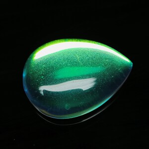 Puede incluir: Una gema iridiscente en forma de lágrima, con un tono verde vibrante. La superficie pulida refleja la luz, creando un efecto brillante. La gema se encuentra sobre un fondo negro, resaltando su color y forma.