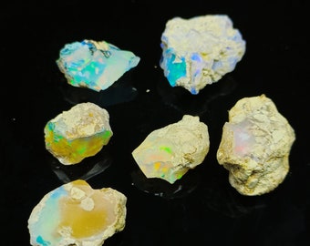 May include: Six rough opal stones with a variety of colors, including white, blue, green, and yellow. The stones are set against a black background.