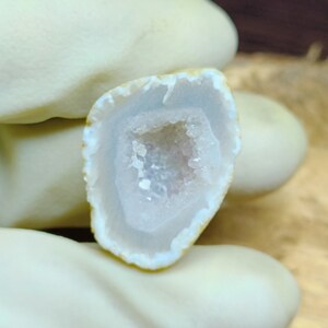 May include: A polished, open geode with a light purple interior and a crystalline center. The outer edge is a light tan color. The geode is held by a gloved hand.