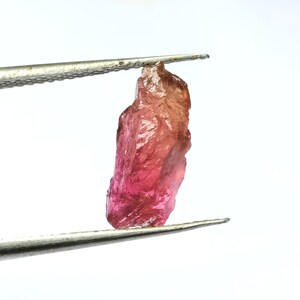 Könnte beinhalten: Ein roher rosafarbener und roter Edelstein, der mit einer Pinzette gehalten wird. Der Edelstein ist ein roher, ungeschliffener Kristall mit einer spitzen Spitze.