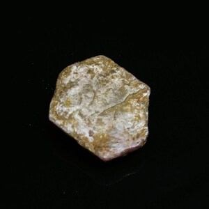 Puede incluir: Una pequeña roca de forma irregular con una superficie rugosa y texturizada. La roca muestra una mezcla de colores, incluyendo beige, marrón y blanco, con una apariencia ligeramente cristalina. El objeto está sobre un fondo negro.