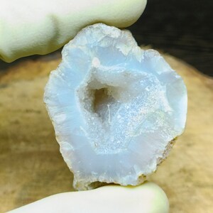 May include: A light blue geode with a hollow center, showcasing a crystalline interior. The outer edges are a light tan color. The geode is held by a gloved hand.