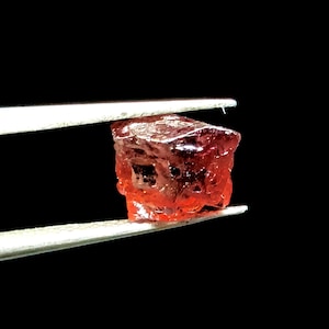 May include: A deep red, faceted gemstone held in tweezers. The gemstone is a rough cut and has a translucent appearance.