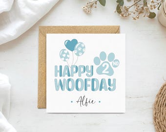 May include: A square greeting card with a white background featuring light blue text that reads Happy 2nd Woofday and the name Alfie. The design includes three balloons, one heart-shaped and two round with paw prints, and a large paw print icon.