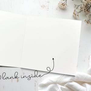 以下が含まれることがあります： 筆記体の「Blank inside」という言葉が入った、開いた状態の白いグリーティングカード。カードは白く、シンプルでエレガントなデザインです。小さな矢印がカードの内側を指しています。カードは白い表面にあります。