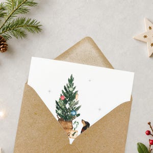 May include: A white greeting card featuring a watercolor illustration of a decorated Christmas tree in a woven basket, a small gift box, a candy cane, and a black and tan dachshund dog, all set inside a brown kraft paper envelope.