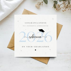 以下が含まれることがあります： 「CONGRATULATIONS TO A WONDERFUL GODDAUGHTER」と「2026 rebecca ON YOUR GRADUATION」の文字が入った卒業祝いのカード。数字は水色で、名前は筆記体。卒業帽子のグラフィックが上にあります。