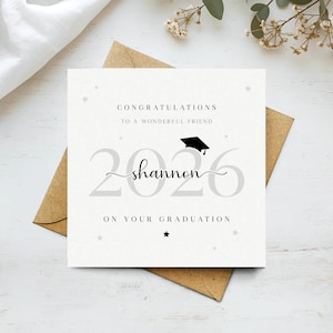 以下が含まれることがあります： 「CONGRATULATIONS TO A WONDERFUL FRIEND」と「2026 shannon ON YOUR GRADUATION」の文字が入った卒業祝いのカード。卒業帽子のアイコンと星のアクセントが特徴です。茶色の封筒の上に置かれています。