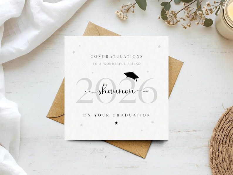 以下が含まれることがあります： 「CONGRATULATIONS TO A WONDERFUL FRIEND」と「2026 shannon ON YOUR GRADUATION」の文字が入った卒業祝いのカード。卒業帽子のアイコンと星のアクセントが特徴です。茶色の封筒の上に置かれています。