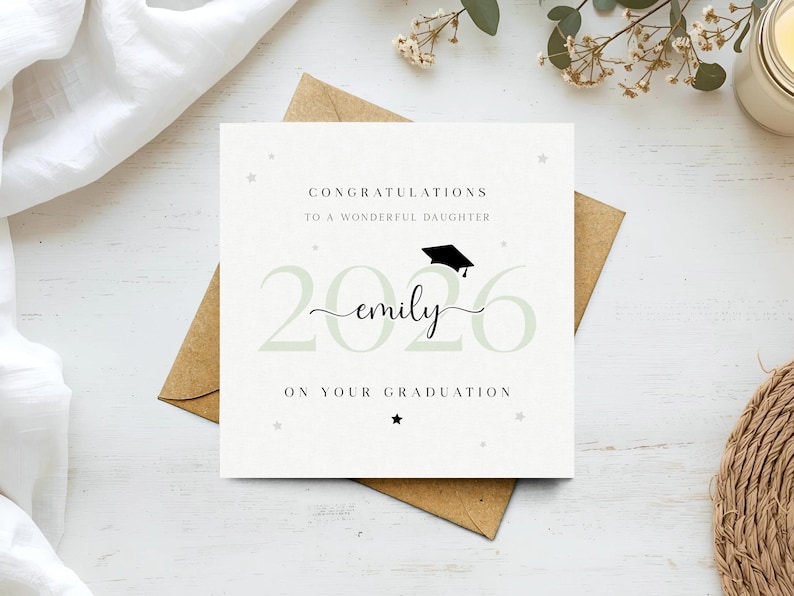 以下が含まれることがあります： 「CONGRATULATIONS TO A WONDERFUL DAUGHTER」と「2026 emily ON YOUR GRADUATION」の文字が入った卒業祝いのカード。卒業帽子のアイコンと星が描かれています。茶色の封筒と白い表面に置かれています。