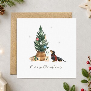 May include: A square greeting card featuring a watercolor illustration of a dachshund wearing a red scarf, sitting beside a decorated Christmas tree in a woven basket with wrapped gifts. The text Merry Christmas is printed at the bottom.