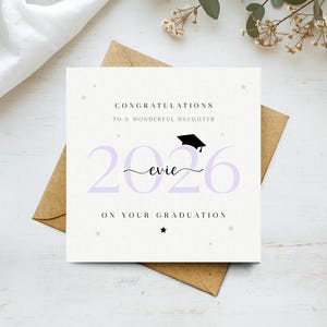 以下が含まれることがあります： 「素晴らしい娘へおめでとう」と「2026 Evie 卒業おめでとう」というテキストが書かれた卒業カード。カードは白地で、ラベンダー色の文字と黒い卒業帽子のイラストが描かれています。茶色の封筒の上に置かれています。