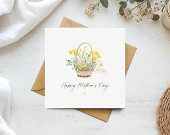 May include: A square greeting card with a watercolor illustration of a basket of yellow and white flowers. The card reads "Happy Mother's Day" in black script, with a small yellow heart. A brown envelope is beneath the card.