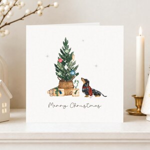 May include: A square greeting card featuring a watercolor illustration of a dachshund wearing a red scarf, standing next to a small Christmas tree in a woven basket with wrapped gifts and the text Merry Christmas below.