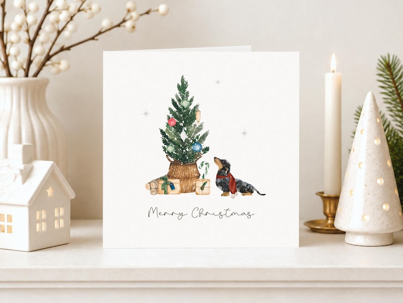 May include: A square greeting card featuring a watercolor illustration of a dachshund wearing a red scarf, standing next to a small Christmas tree in a woven basket with wrapped gifts and the text Merry Christmas below.