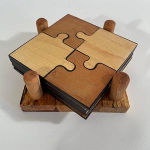 Puede incluir: Posavasos de madera con forma de piezas de rompecabezas, en tonos marrón y beige, apilados sobre un soporte de madera. Los posavasos están diseñados para encajar como un rompecabezas. El soporte tiene cuatro clavijas de madera.