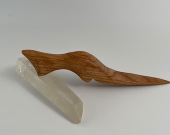 May include: A wooden hair stick with a curved design and a pointed end, resting on a clear quartz crystal. The hair accessory is made of light brown wood and is set against a white background.
