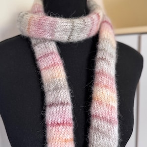 May include: A long, knitted scarf featuring a soft, fuzzy texture with a variegated colour pattern of light pink, peach, cream, and soft grey stripes. The scarf is wrapped around a black mannequin neck, displaying a consistent stockinette stitch.