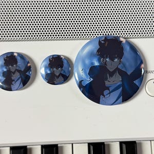 May include: Three round buttons with a blue and white background featuring a black and white anime character with black hair. The character is wearing a black jacket and a white shirt. The buttons are different sizes.