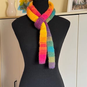 May include: A rainbow-coloured knitted scarf draped on a black mannequin. The scarf showcases a vibrant gradient of colours, including pink, orange, yellow, green, and purple. The scarf is made of a soft, textured yarn, ideal for adding a pop of colour to any outfit.