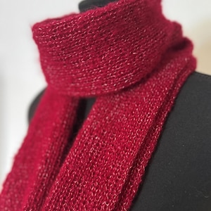 May include: A knitted red scarf with a subtle shimmer. The scarf is draped over a black form, highlighting its texture and design. The close-up shot emphasises the intricate knit pattern and the rich colour of the scarf.
