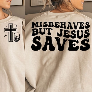 May include: A beige sweatshirt with the text "Misbehaves But Jesus Saves" printed in black on the back. The front of the sweatshirt has a black cross with a small hand gesture graphic.