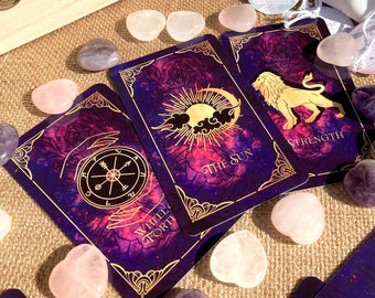 Peut inclure: Un ensemble de cartes de tarot, de cristaux et de petits sacs à cordon. Les cartes de tarot présentent des illustrations dorées et les mots "Wheel of Fortune", "The Sun" et "Strength". Les cristaux sont roses, violets et transparents. Les sacs ont des motifs imprimés.