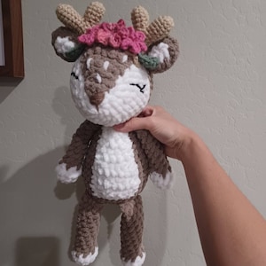 May include: A crocheted deer plush toy with brown and white fur, wearing a pink flower crown and green leaves.
